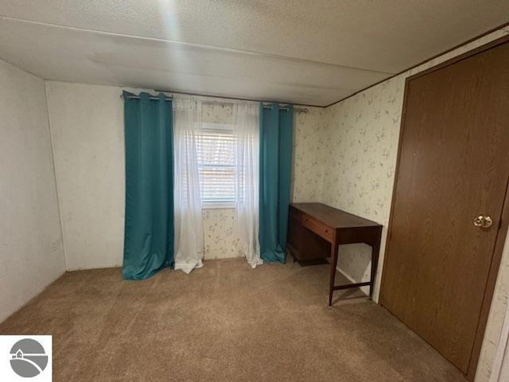 Interior view of a well-lit room featuring a window with sheer and teal curtains, beige carpeting, a wooden desk, and a closed wooden door, reflecting the cozy atmosphere of the 3-bedroom home for sale at 2520 S Blodgett, Lake City, MI.