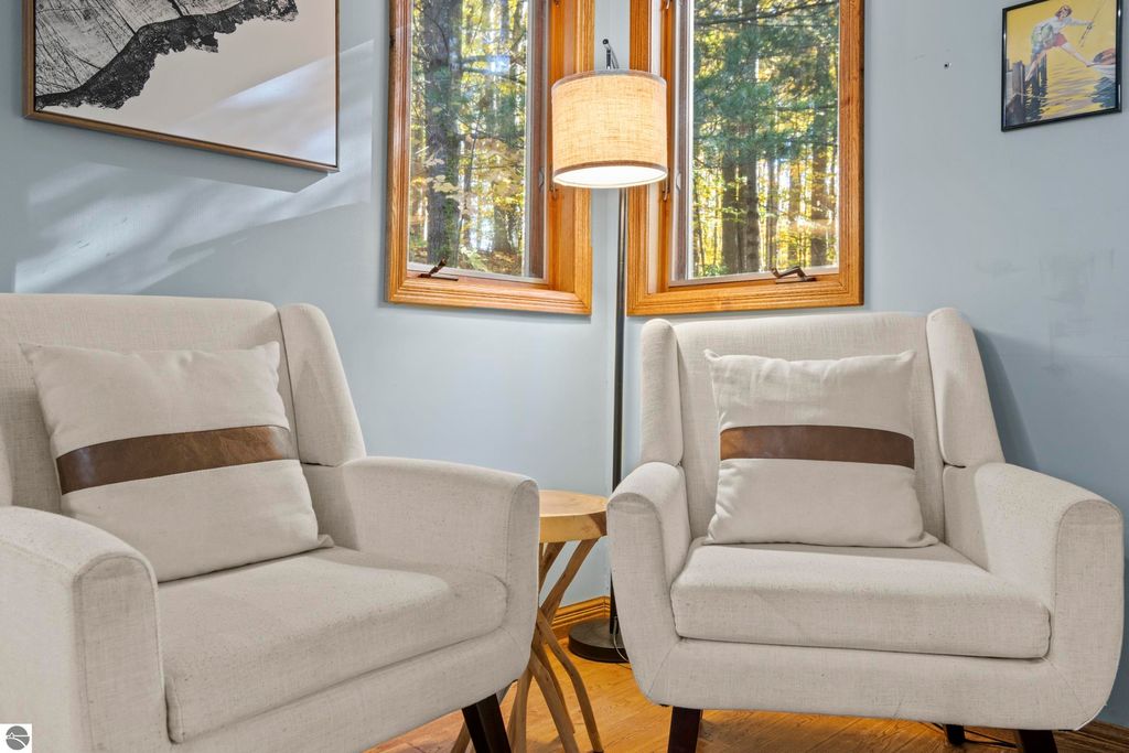 Cozy living space featuring two modern armchairs with decorative pillows, a wooden side table, and large windows showcasing a serene wooded view, highlighting the home’s connection to nature at 10371 Deerpath North, Traverse City, MI.