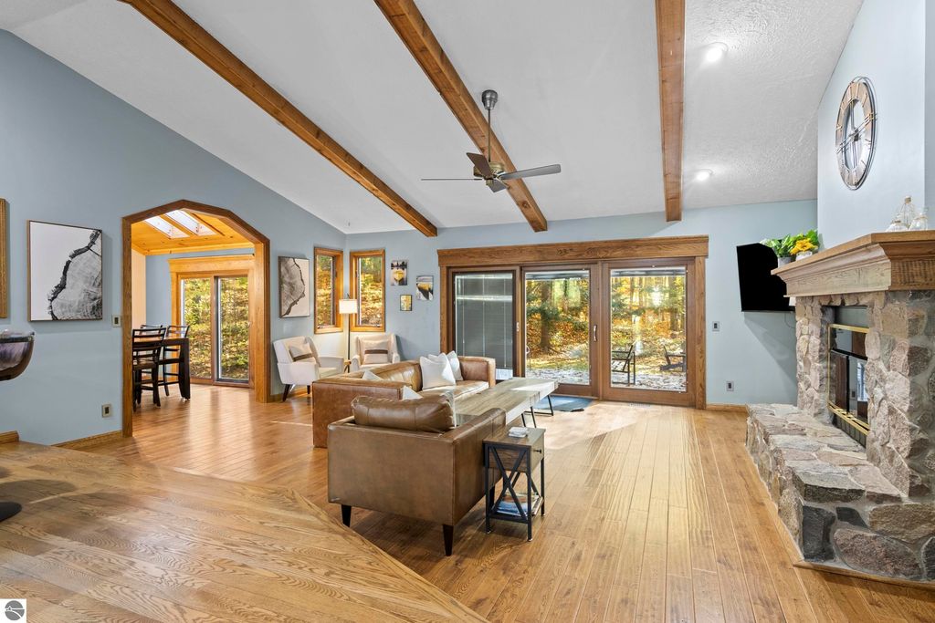 Interior view of a spacious living room featuring wooden beams, a stone fireplace, and large windows overlooking a wooded area, showcasing a blend of comfort and natural light in a luxury home at 10371 Deerpath North, Traverse City, MI.