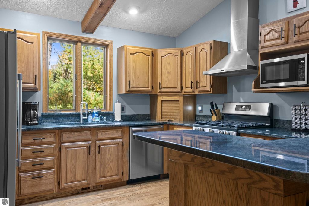 Modern kitchen with oak cabinetry, granite countertops, stainless steel appliances, and large windows showcasing a wooded view, highlighting the home's connection to nature at 10371 Deerpath North, Traverse City.