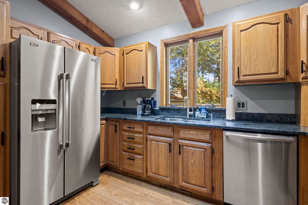Modern kitchen featuring stainless steel refrigerator, wooden cabinets, granite countertops, and large windows with wooded view, highlighting the spacious layout of the home at 10371 Deerpath North, Traverse City, MI.