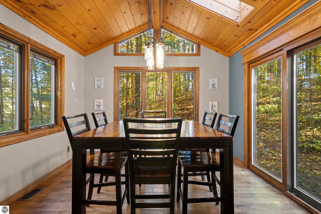 Dining area with large wooden table, surrounded by windows showcasing wooded views, featuring a wooden ceiling and contemporary light fixtures, emphasizing the home's connection to nature.