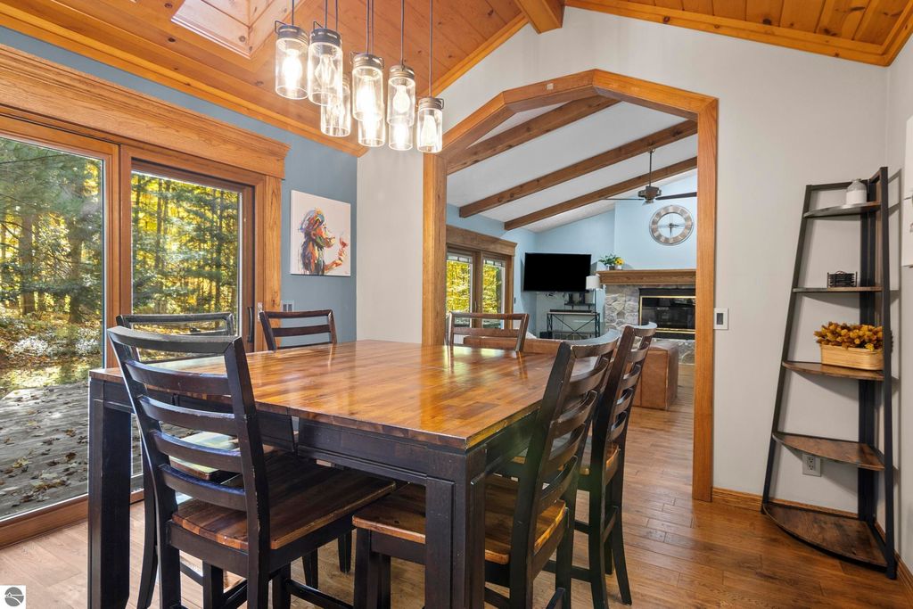 Dining area with wooden table and chairs, large windows showcasing wooded views, modern light fixtures, and open layout leading to living space with fireplace and TV, reflecting the luxury and comfort of the home at 10371 Deerpath North, Traverse City, MI.
