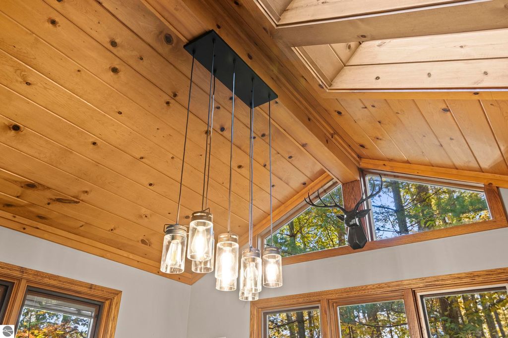 Ceiling with wooden beams, modern pendant lights, and a mounted deer head, featuring large windows that showcase wooded surroundings, enhancing the rustic luxury of the Traverse City home.