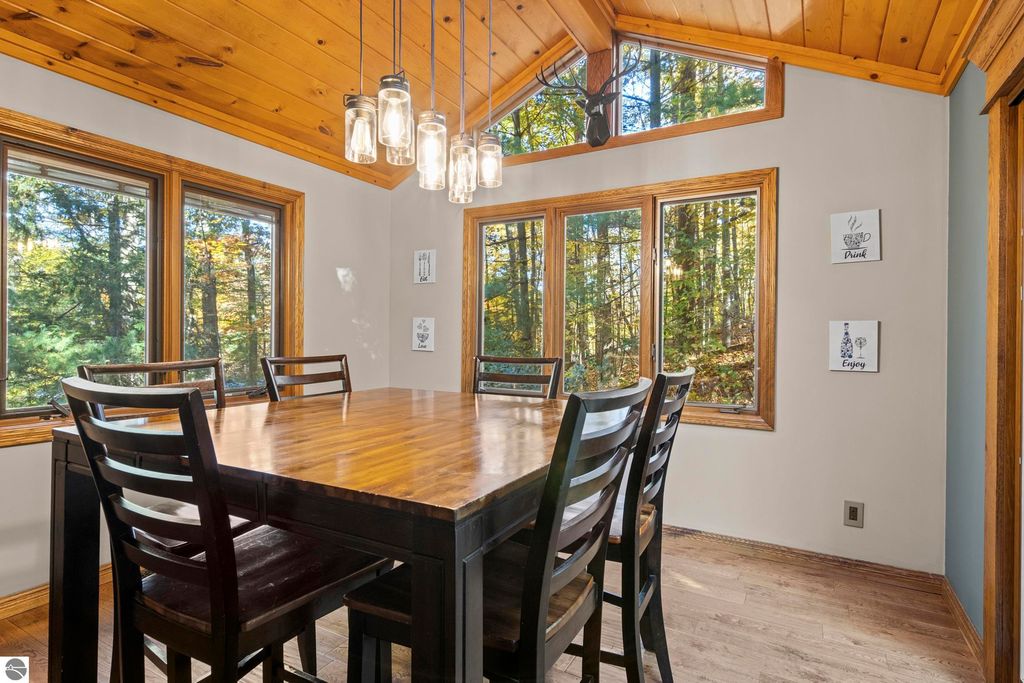 Dining area with a wooden table, chairs, and large windows showcasing wooded surroundings, featuring pendant lighting and decorative wall art, highlighting the home's connection to nature at 10371 Deerpath North, Traverse City, MI.