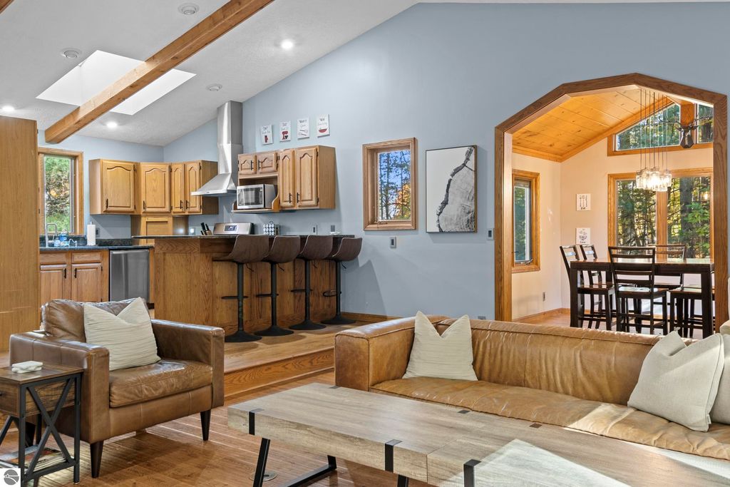 Modern living room interior with leather sofas, wooden accents, and a kitchen featuring stainless steel appliances, granite countertops, and a breakfast bar, showcasing a spacious layout and natural light from large windows.