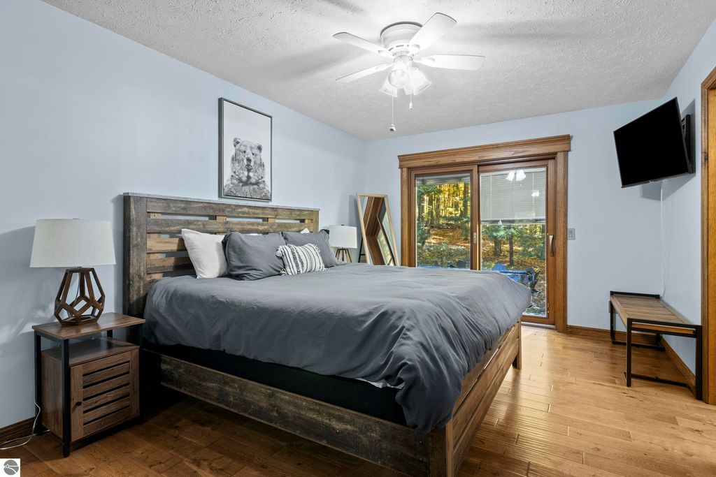 Bedroom featuring a wooden bed frame with gray bedding, bedside tables with lamps, a bear-themed wall art, and large windows overlooking a wooded area in a luxury home at 10371 Deerpath North, Traverse City, MI.
