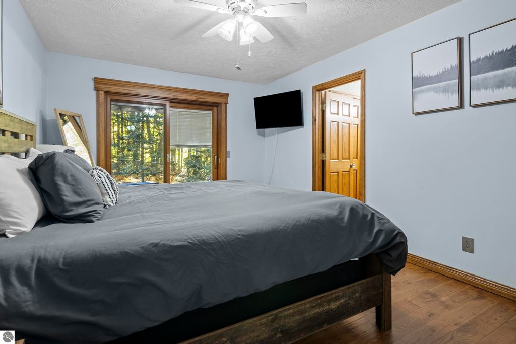 Cozy bedroom featuring a large bed with gray bedding, wooden accents, a mounted TV, and large windows overlooking a wooded area, complemented by nature-themed artwork on the walls.