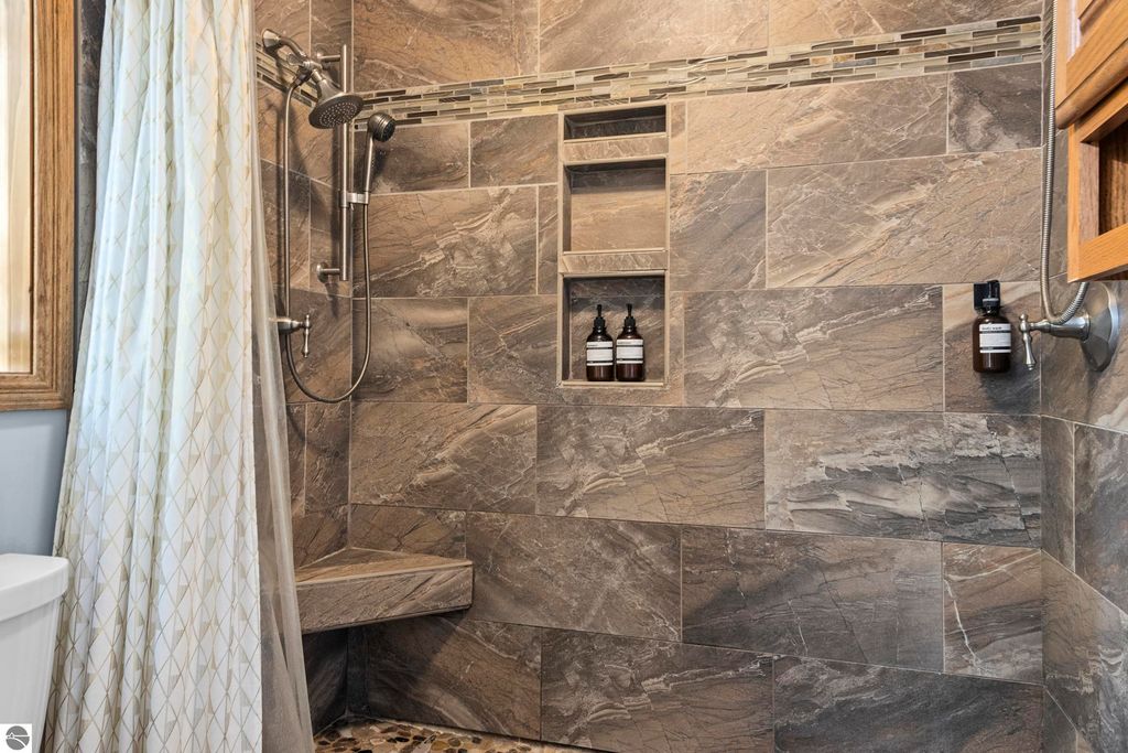 Luxury bathroom shower with modern fixtures, stone tiles, built-in shelves for toiletries, and a glass shower curtain, showcasing elegant design in the Traverse City home listing.