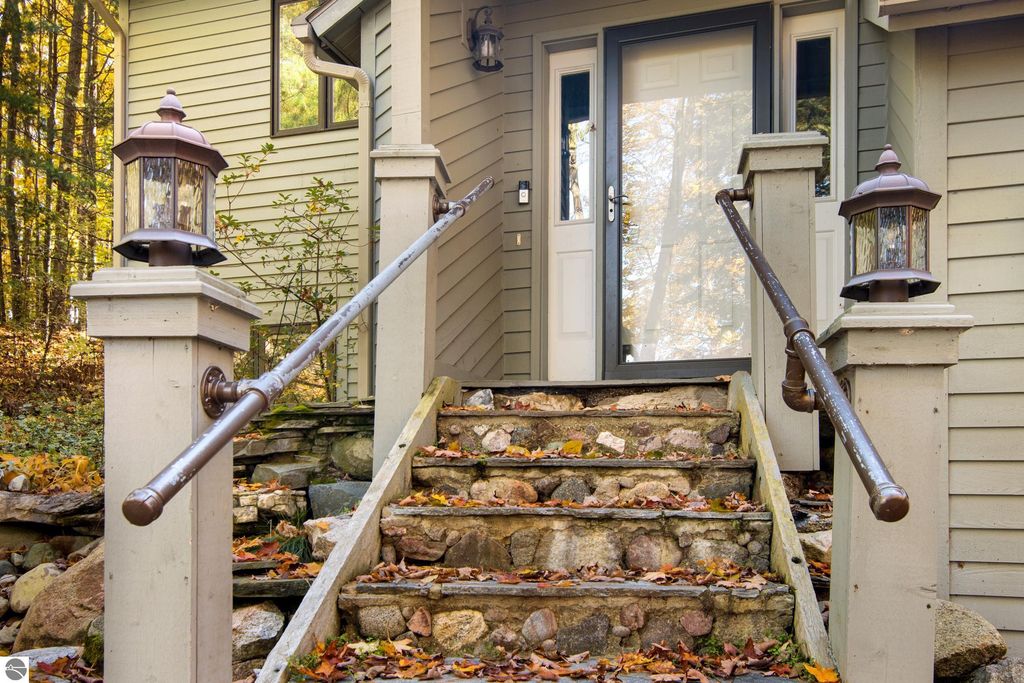 Front entrance of 10371 Deerpath North featuring stone steps, decorative lanterns, and a modern door, surrounded by autumn foliage, emphasizing the home's connection to nature and luxury living.