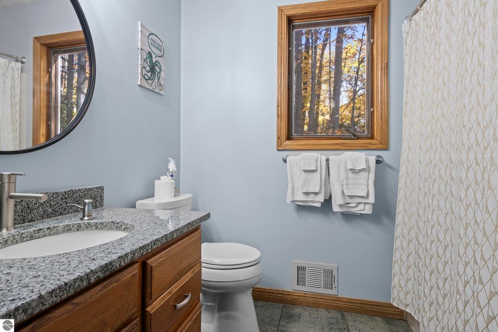 Bathroom with granite countertop, round mirror, and window showing wooded view, featuring towels and modern fixtures, reflecting luxury home amenities at 10371 Deerpath North, Traverse City.