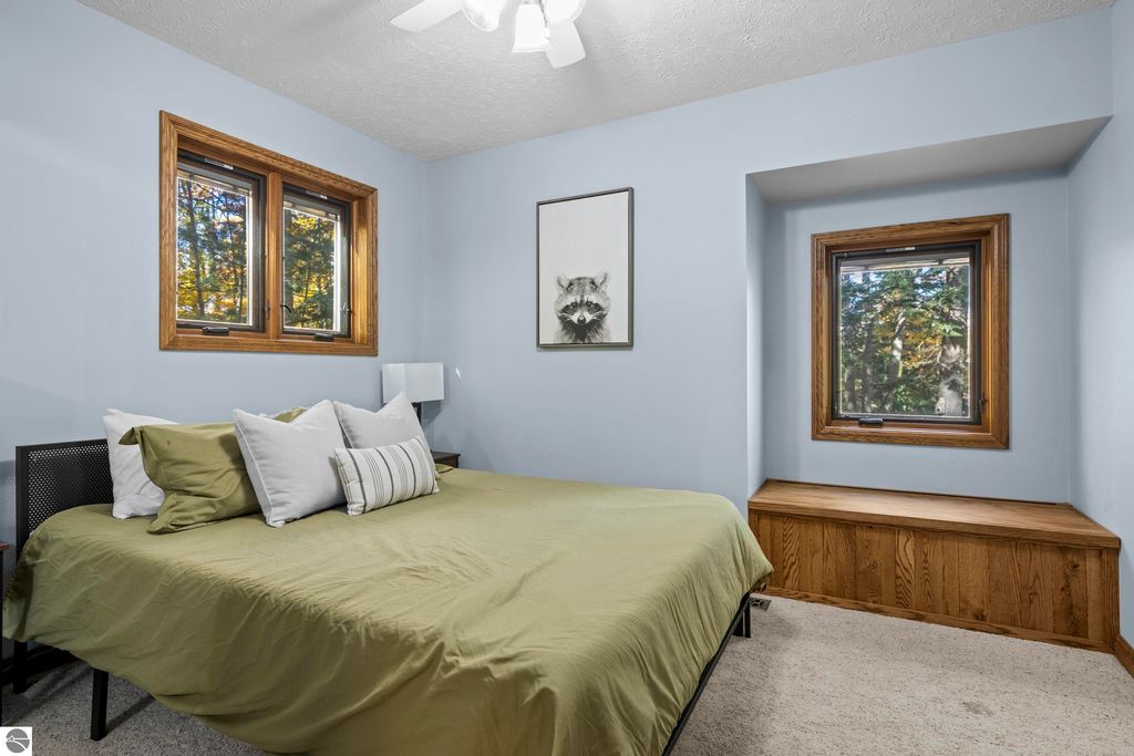 Cozy bedroom with green bedding, decorative pillows, large windows showcasing wooded views, and a framed raccoon artwork on the wall, highlighting the serene atmosphere of the Traverse City home.