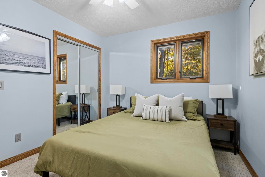 Cozy bedroom featuring a green bedspread, decorative pillows, and bedside tables with lamps, complemented by large windows showcasing wooded views, reflecting the serene nature of the 10371 Deerpath North property in Traverse City, MI.