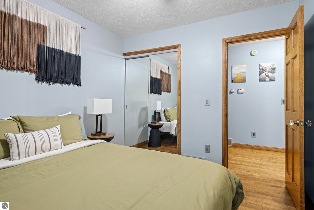 Cozy bedroom featuring a green bedspread, decorative pillows, a bedside lamp, and wall art, with a door leading to another room, reflecting the stylish updates of the Traverse City property listing at 10371 Deerpath North.