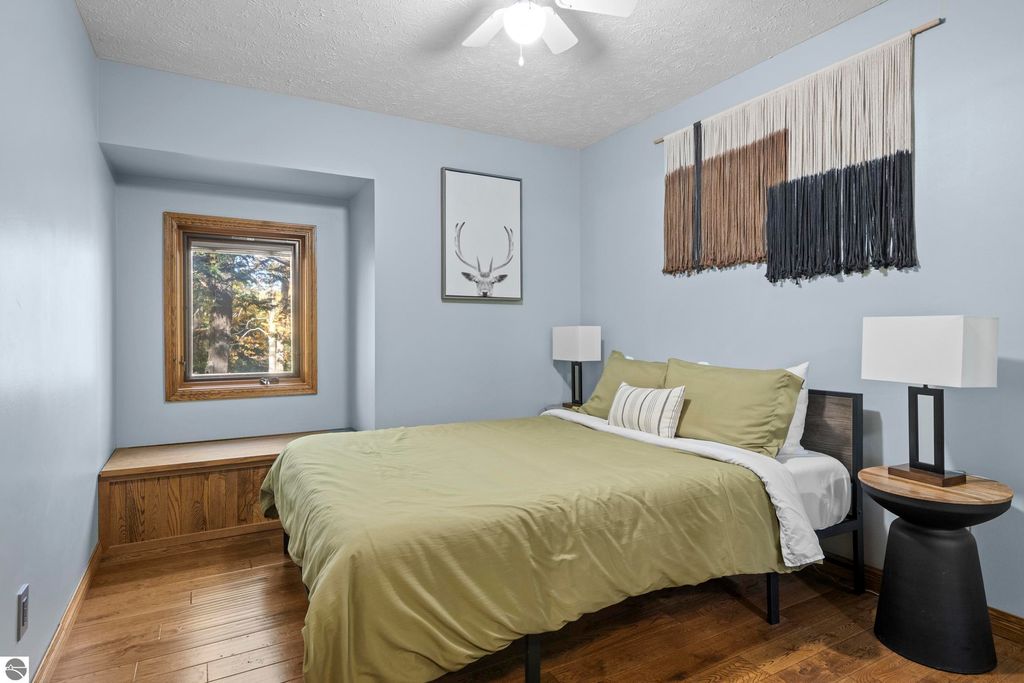 Cozy bedroom featuring a green bedspread, wooden accents, a window with forest views, and modern decor, part of the luxurious home at 10371 Deerpath North, Traverse City.