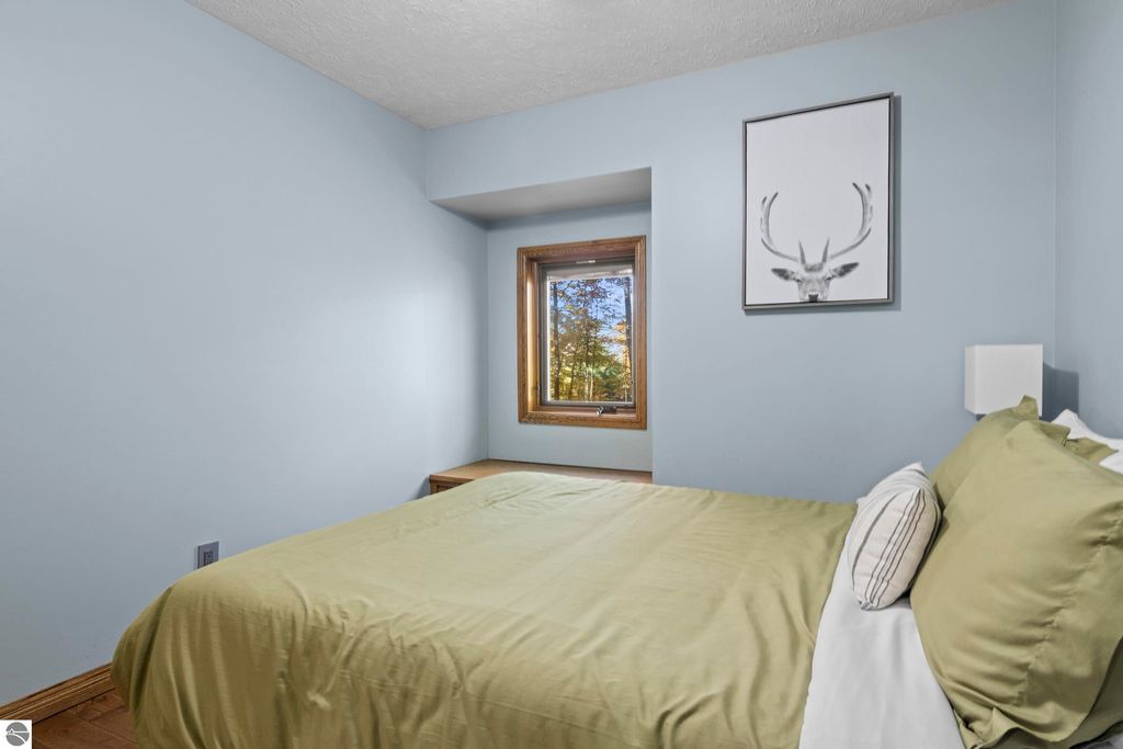 Cozy bedroom with a green bedspread, wooden accents, and a window showcasing wooded views, featuring a framed deer antler artwork on the wall.