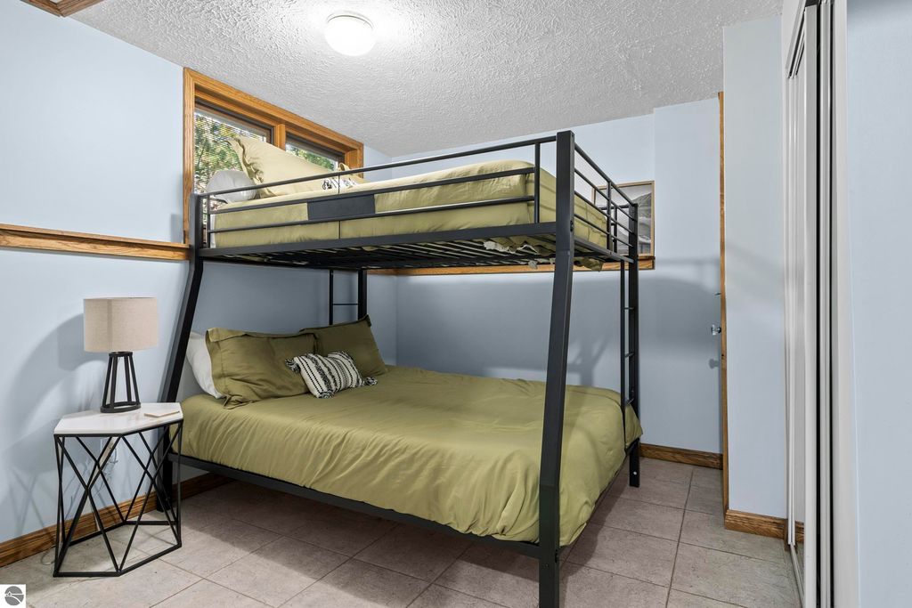 Cozy bedroom with a bunk bed and a double bed, featuring light blue walls, a bedside table with a lamp, and natural light from a window, highlighting the comfortable and inviting atmosphere of the property at 10371 Deerpath North in Traverse City, MI.