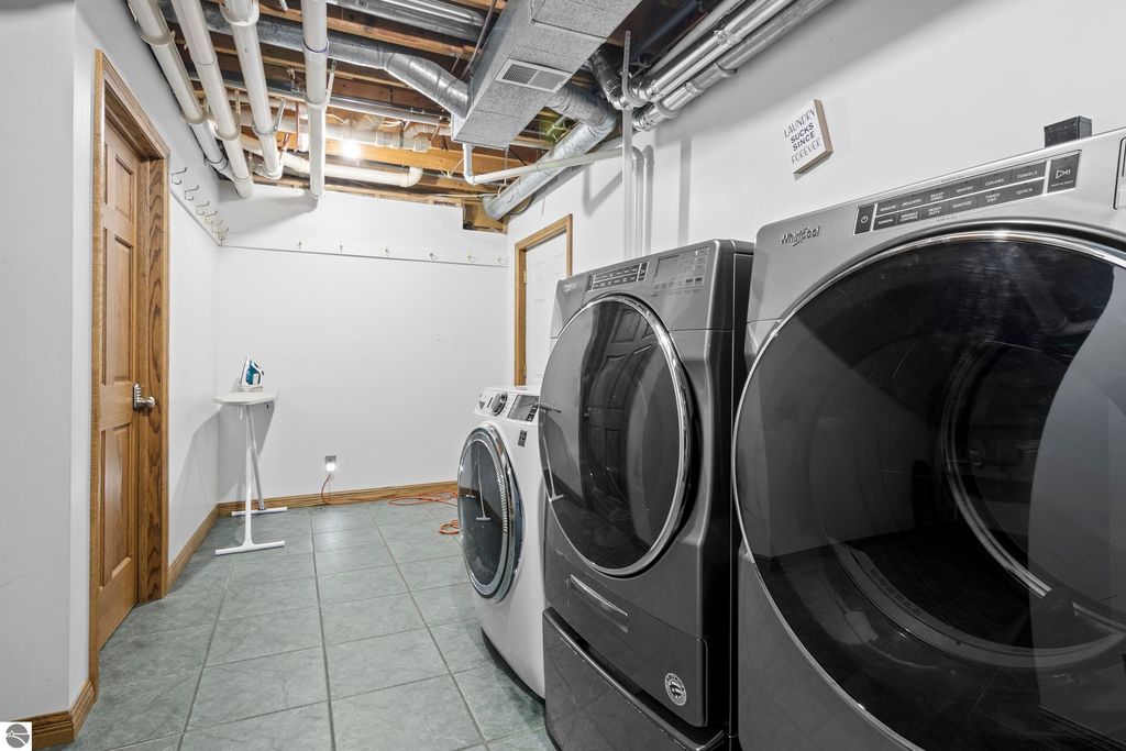 Laundry room featuring modern washer and dryer, tiled floor, and ironing board, with a door leading to another space and exposed pipes above.