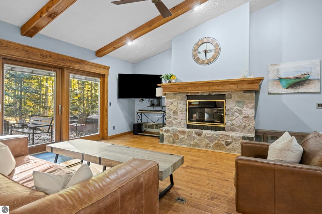 Cozy living room with stone fireplace, large windows showcasing wooded views, leather furniture, and modern decor at 10371 Deerpath North, Traverse City, MI.