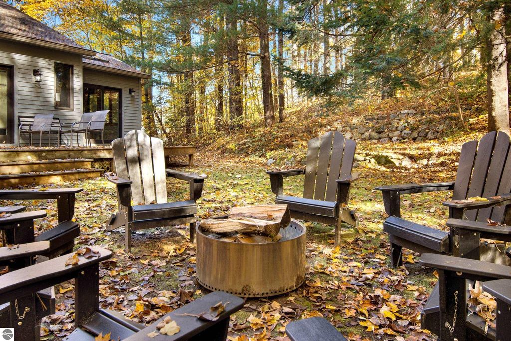 Outdoor seating area with Adirondack chairs surrounding a fire pit, set in a wooded landscape with autumn foliage, adjacent to a modern home in Traverse City, MI, highlighting the property’s connection to nature and outdoor living.