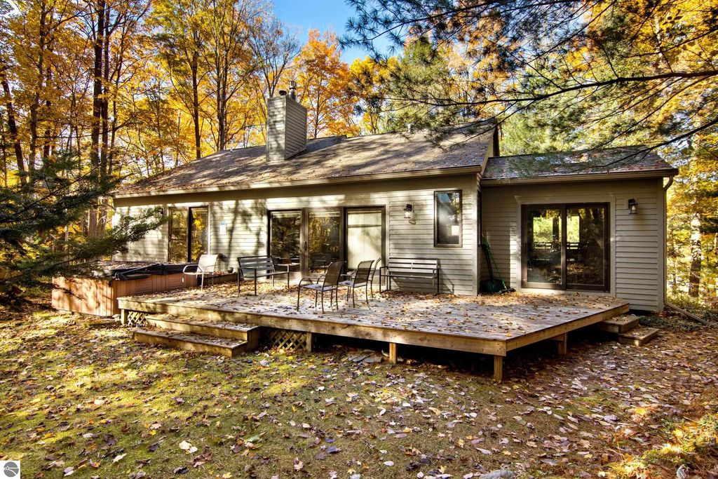 Exterior view of a modern home at 10371 Deerpath North, Traverse City, featuring a spacious wooden deck, hot tub, and surrounded by colorful autumn foliage, highlighting the serene wooded setting and luxury lake access.