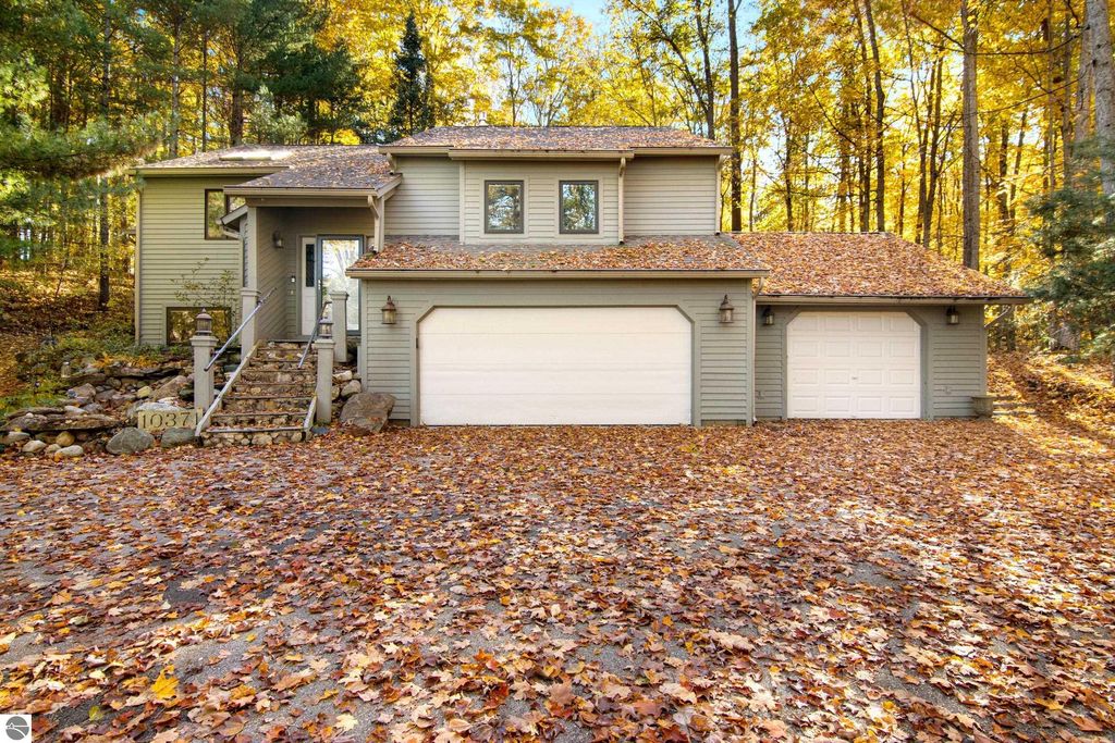 Exterior view of 10371 Deerpath North, Traverse City, MI, showcasing a modern home surrounded by colorful autumn foliage, featuring a two-car garage and stone pathway leading to the entrance.