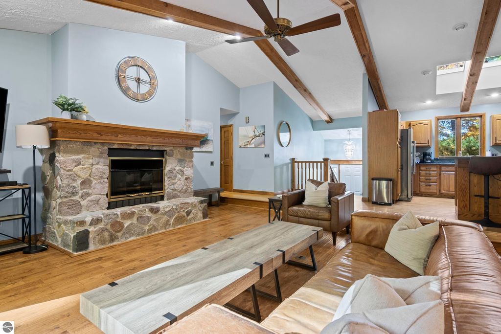 Cozy living room featuring a stone fireplace, wooden beams, modern furniture, and an open layout leading to a kitchen, showcasing a blend of rustic charm and contemporary design.